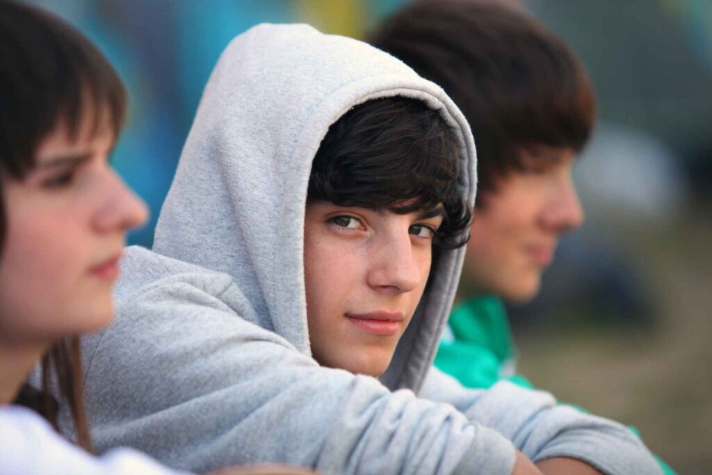 Three teenage boys sitting together outdoors, with one wearing a gray hoodie and looking toward the camera.